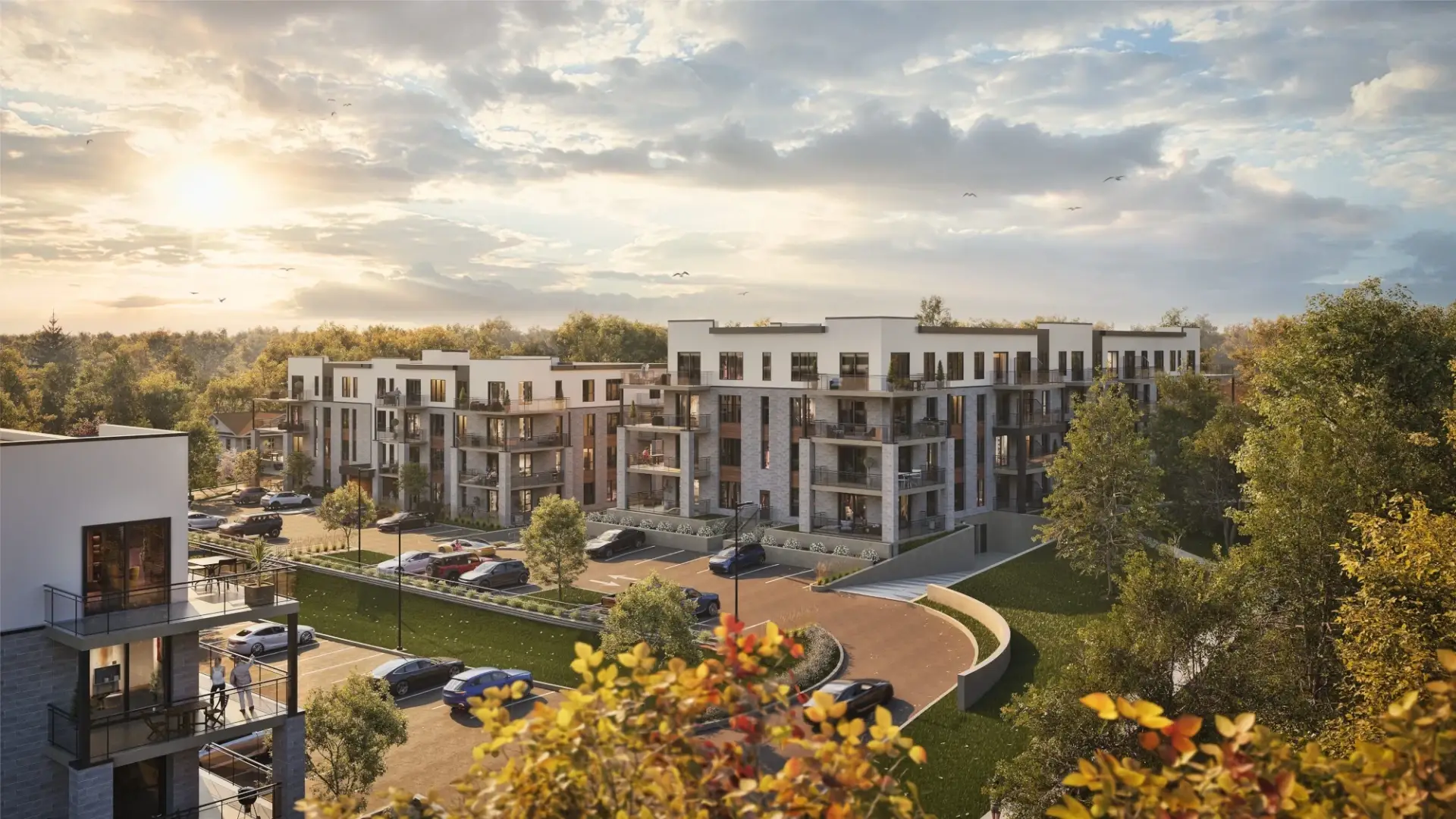 Des immeubles d'habitation modernes entourés d'arbres et de verdure, avec des voitures garées devant et le soleil se couchant dans un ciel partiellement nuageux, créant une atmosphère chaleureuse et invitante.